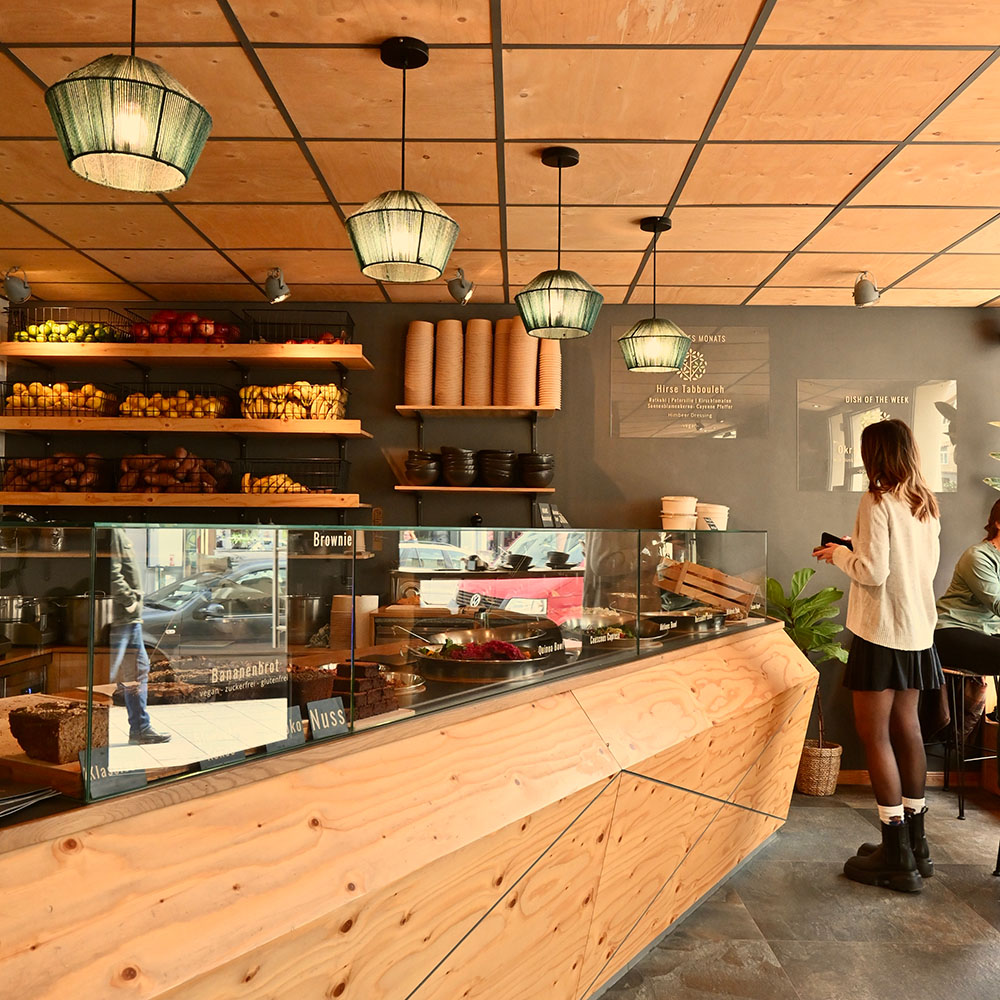 Kundin steht vor einer Restauranttheke mit verschiedenen Speisen wie Quinoa Bowl und Couscous Caprese. Im Hintergrund befinden sich Regale mit frischem Obst und Verpackungen. Die Theke ist aus Holz, und moderne Lampen hängen von der Decke.