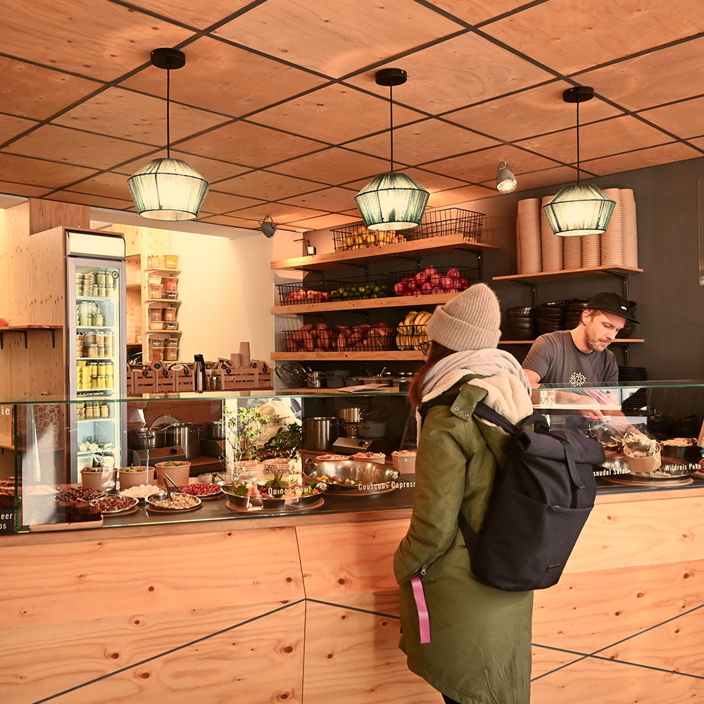 Kundin mit Rucksack steht an der Restauranttheke, hinter der verschiedene Gerichte wie Quinoa Bowl und Couscous Caprese ausgestellt sind. Ein Mitarbeiter bereitet Speisen hinter der Theke vor. Im Hintergrund Regale mit frischem Obst und Verpackungen.