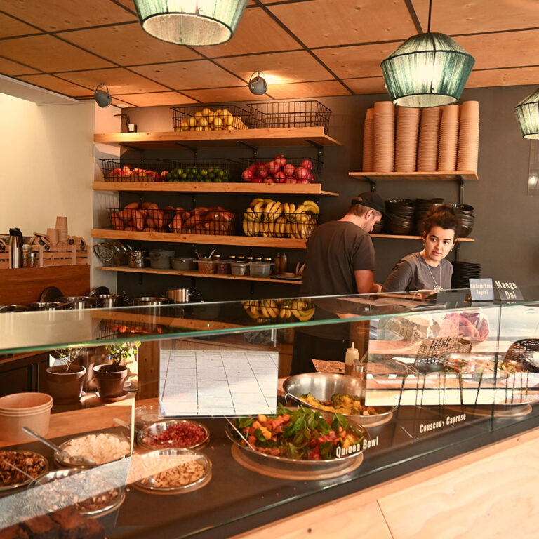 Restauranttheke mit frischem Obst, Salaten und Quinoa Bowl, zwei Mitarbeiter hinter der Theke bei der Zubereitung.